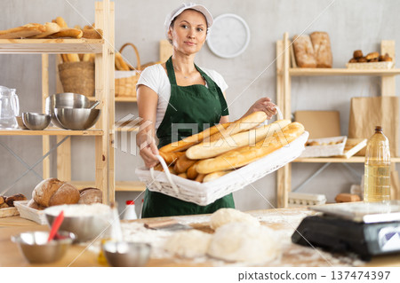 Positive young woman baker in uniform baking fresh baguettes in bakery Positive young woman baker in uniform baking fresh baguettes in bakery 137474397