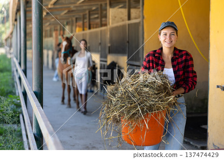 European young female worker walks go through corridor of stable, carrying basket with straw 137474429