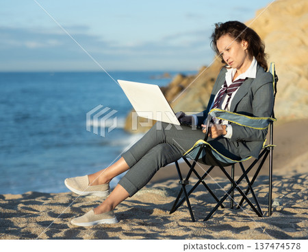 Businesswoman in office clothes working on laptop on seashore Businesswoman in office clothes working on laptop on seashore 137474578