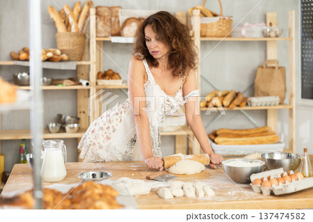 Woman in sundress is standing in kitchen of bakery and kneading dough, working with rolling pin Woman in sundress is standing in kitchen of bakery and kneading dough, working with rolling pin 137474582