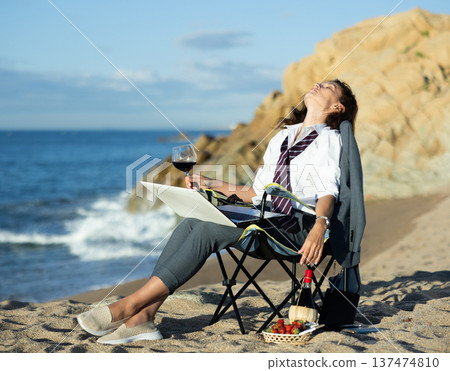 Stylish woman in a suit works on her laptop by the ocean and drinking wine Stylish woman in a suit works on her laptop by the ocean and drinking wine 137474810