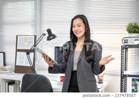 Happy businesswoman or office worker holding tablet open both arms while standing at computer table. 137478587