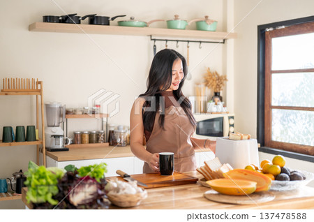 Asian woman holding coffee waiting for bread in toaster aside vegetables on kitchen cooking counter. Asian woman holding coffee waiting for bread in toaster aside vegetables on kitchen cooking counter. 137478588