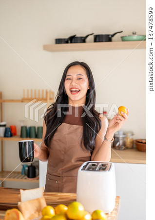 Pretty asian woman holding coffee mug and orange standing aside toaster on kitchen cooking counter. Pretty asian woman holding coffee mug and orange standing aside toaster on kitchen cooking counter. 137478599