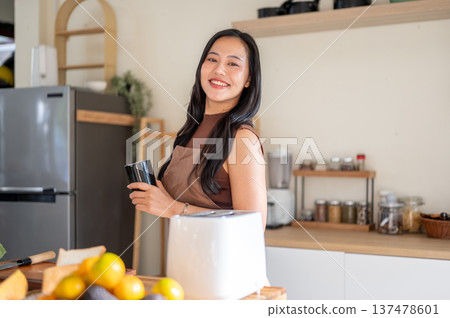Pretty asian woman holding a coffee mug while standing aside toaster and fruits on kitchen counter. Pretty asian woman holding a coffee mug while standing aside toaster and fruits on kitchen counter. 137478601