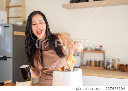 Pretty asian woman holding coffee mug while excitedly taking bread from toaster on kitchen counter. Pretty asian woman holding coffee mug while excitedly taking bread from toaster on kitchen counter. 137478604