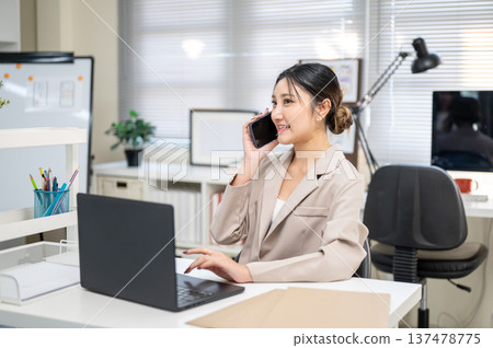 Asian businesswoman office worker holding phone talking on call sitting in front of laptop on table. Asian businesswoman office worker holding phone talking on call sitting in front of laptop on table. 137478775