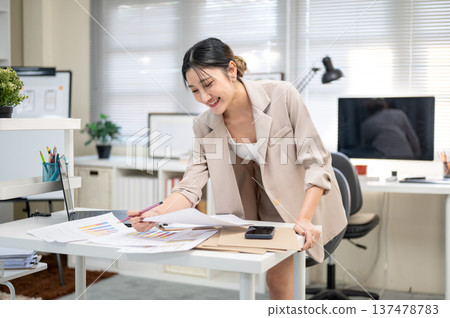 Asian businesswoman or office worker holding pencil looking at data on document as standing at table Asian businesswoman or office worker holding pencil looking at data on document as standing at table 137478783