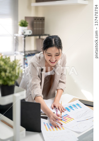 Asian businesswoman or office worker leaning over table holding pencil looking at data in documents. Asian businesswoman or office worker leaning over table holding pencil looking at data in documents. 137478784