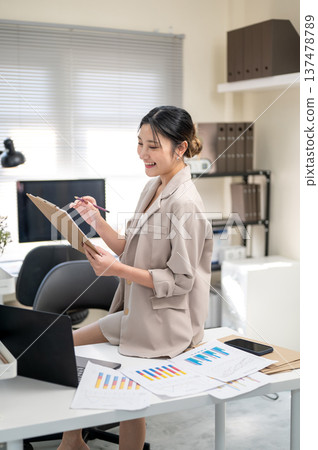 Asian businesswoman or office worker holding pencil looking at clipboard sits on table with document. Asian businesswoman or office worker holding pencil looking at clipboard sits on table with document. 137478789