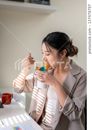 Hungry asian businesswoman or office worker eating instant noodle cup at table with bright sunlight. 137478797