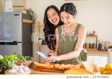 Asian woman laughing as girlfriend holding knife cutting papaya on board at kitchen cooking counter. Asian woman laughing as girlfriend holding knife cutting papaya on board at kitchen cooking counter. 137478962