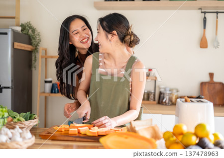 Asian woman smiling at girlfriend holding knife cutting papaya on board at kitchen cooking counter. 137478963