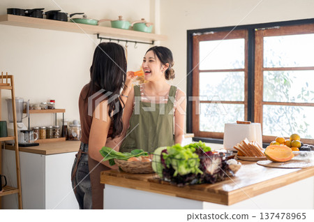 Asian woman feeding fruit to girlfriend as standing at kitchen cooking counter with food ingredients Asian woman feeding fruit to girlfriend as standing at kitchen cooking counter with food ingredients 137478965