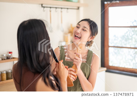 Happy asian woman couple girlfriends holding wooden spatula singing having fun in a house's kitchen. Happy asian woman couple girlfriends holding wooden spatula singing having fun in a house's kitchen. 137478971