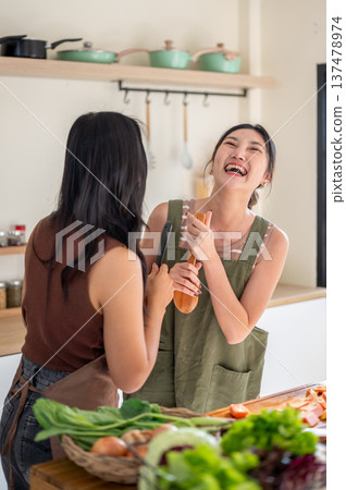 Asian woman holding spatula laughing at girlfriend at kitchen counter while cooking food ingredients Asian woman holding spatula laughing at girlfriend at kitchen counter while cooking food ingredients 137478974
