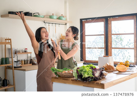 Asian woman holding spatula enjoy singing to a girlfriend at kitchen counter while cooking together. Asian woman holding spatula enjoy singing to a girlfriend at kitchen counter while cooking together. 137478976