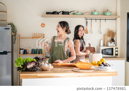 Pretty asian woman throwing onion having fun aside girlfriend while cooking food at kitchen counter. 137478981