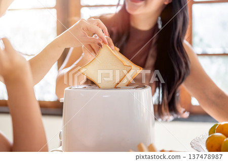 Close up of a woman looking at girlfriend while putting bread in toaster standing at kitchen counter Close up of a woman looking at girlfriend while putting bread in toaster standing at kitchen counter 137478987