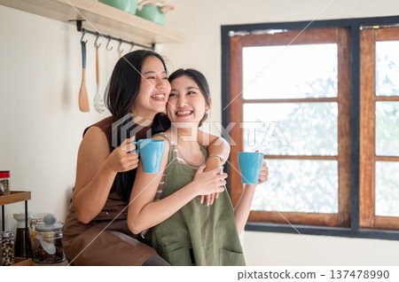 Happy asian woman girlfriends holding couple mug drinking coffee while hugging at kitchen counter. 137478990