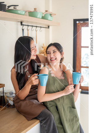 Happy asian woman girlfriends holding couple mug drinking coffee while cuddling at kitchen counter. Happy asian woman girlfriends holding couple mug drinking coffee while cuddling at kitchen counter. 137478991