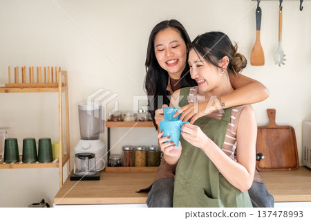 Pretty asian woman girlfriends holding couple mug drinking coffee while hugging at kitchen counter. 137478993