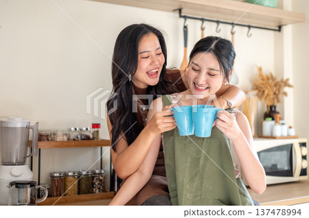 Happy asian woman girlfriends clinking couple mugs drinking coffee while hugging at kitchen counter. 137478994