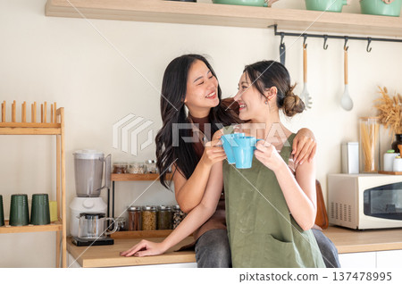 Smiling asian woman girlfriends clinking couple mug drinking coffee while hugging at kitchen counter 137478995