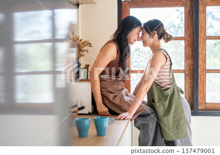 Pretty asian woman cornering touching foreheads with her girlfriend who sitting on kitchen counter. Pretty asian woman cornering touching foreheads with her girlfriend who sitting on kitchen counter. 137479001
