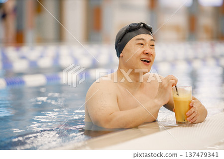 Man wearing special hat and goggles for swimming. Asian man standing in water in swimming pool and drinking an orange juice. Professionale male swimmer has a break. 137479341