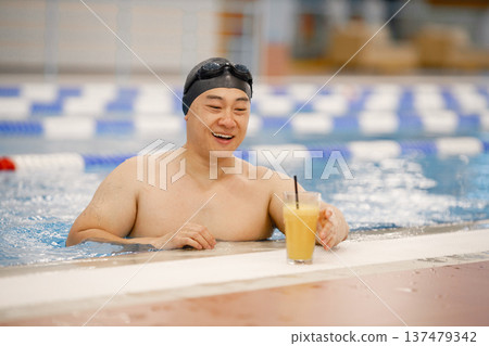 Man wearing special hat and goggles for swimming. Asian man standing in water in swimming pool and drinking an orange juice. Professionale male swimmer has a break. Man wearing special hat and goggles for swimming. Asian man standing in water in swimming pool and drinking an orange juice. Professionale male swimmer has a break. 137479342