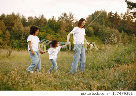 Photo of caucasian mother and two her african american daughters walking together outdoors. Girls and mother holding each others hands. Mother and daughers wearing white t-shirts. Photo of caucasian mother and two her african american daughters walking together outdoors. Girls and mother holding each others hands. Mother and daughers wearing white t-shirts. 137479391