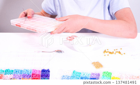 Delicate fingers of a young girl navigate through a treasure trove of bright, multicolored beads, each compartment revealing a new hue to choose from. 137481491
