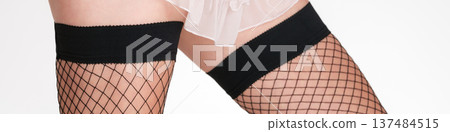 Sensual close-up of a woman's legs in black fishnet stockings with thick bands. Studio shot on clean background, highlighting fashion, allure and texture. 137484515