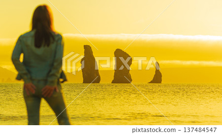 A blurred woman from behind admires a serene ocean view with silhouetted sea stacks. The entire landscape is drenched in a golden yellow light, evoking a peaceful, travel mood. Focus on background 137484547