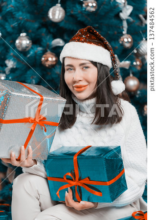 Woman in Santa hat and white sweater holds silver and teal Xmas gifts. Festive holiday female portrait with a stylized, color graded teal Christmas tree background. Cheerful celebratory mood Woman in Santa hat and white sweater holds silver and teal Xmas gifts. Festive holiday female portrait with a stylized, color graded teal Christmas tree background. Cheerful celebratory mood 137484562