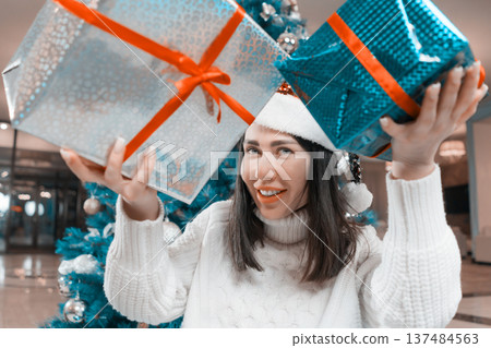 A joyful woman in a white sweater and Santa hat smiles while holding two large Xmas presents. Festive holiday scene with a decorated Christmas tree. Modern teal, red and silver color palette A joyful woman in a white sweater and Santa hat smiles while holding two large Xmas presents. Festive holiday scene with a decorated Christmas tree. Modern teal, red and silver color palette 137484563