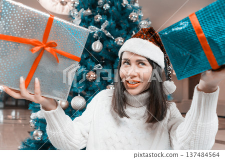 A happy young woman in a white sweater and sparkly red Santa hat smiles holding two Xmas gift boxes. A decorated blue Christmas tree with silver ornaments is in the background. Festive holiday mood A happy young woman in a white sweater and sparkly red Santa hat smiles holding two Xmas gift boxes. A decorated blue Christmas tree with silver ornaments is in the background. Festive holiday mood 137484564