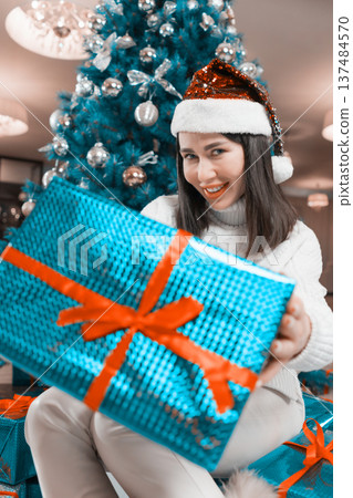 A joyful young woman in a white sweater and red sequined Santa hat smiles as she holds out a large, blue wrapped Xmas gift with a red ribbon. A decorated teal Christmas tree is in the background A joyful young woman in a white sweater and red sequined Santa hat smiles as she holds out a large, blue wrapped Xmas gift with a red ribbon. A decorated teal Christmas tree is in the background 137484570