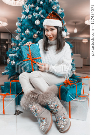Happy woman in a Santa hat sits by a teal Christmas tree, opening a Xmas present. Festive holiday concept with stylish clothes, jeweled boots, and a vibrant red and blue color scheme Happy woman in a Santa hat sits by a teal Christmas tree, opening a Xmas present. Festive holiday concept with stylish clothes, jeweled boots, and a vibrant red and blue color scheme 137484572
