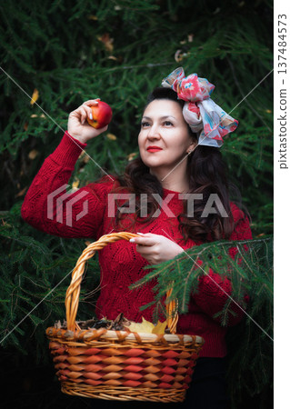 Woman in red sweater holding ripe apple and wicker basket in dark green forest. Portrait with nostalgic, fairytale mood and vintage style. Soft low lighting with red and green color palette Woman in red sweater holding ripe apple and wicker basket in dark green forest. Portrait with nostalgic, fairytale mood and vintage style. Soft low lighting with red and green color palette 137484573