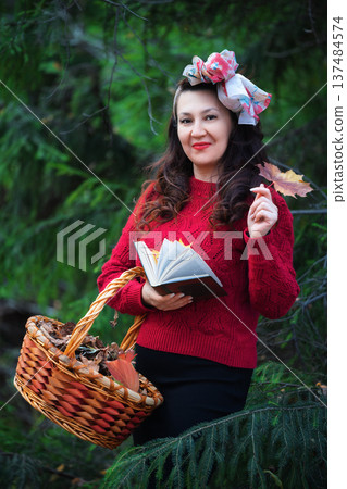 Woman in red knitted sweater smiles and looking at camera in an autumn forest. Happy woman holding book and wicker basket filled with fall leaves. Soft low light creates peaceful, cozy, storybook mood Woman in red knitted sweater smiles and looking at camera in an autumn forest. Happy woman holding book and wicker basket filled with fall leaves. Soft low light creates peaceful, cozy, storybook mood 137484574