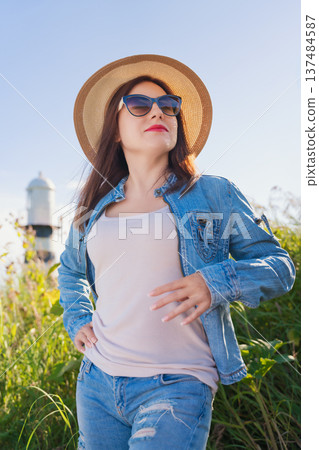 A confident woman in a straw hat, sunglasses, denim jacket enjoys a warm, sunny day. Low-angle shot with a coastal lighthouse in the background. Bright, natural light a blue sky create a happy mood A confident woman in a straw hat, sunglasses, denim jacket enjoys a warm, sunny day. Low-angle shot with a coastal lighthouse in the background. Bright, natural light a blue sky create a happy mood 137484587