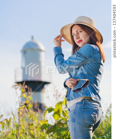 Confident woman in a denim jacket and jeans poses outdoors. Wearing a straw hat, model looking over her shoulder with a blurry lighthouse behind her. Sunny, warm lighting evokes a relaxed travel mood 137484593