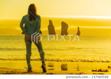 A woman stands on a sandy shore, her figure blurred, looking out at majestic ocean stacks during a brilliant golden sunset. The warm, yellow light creates a dreamy mood. Selective focus on background 137484598