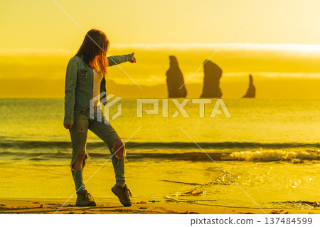 Young woman in ripped jeans stands on a beach, pointing at distant ocean stacks. The scene is bathed in a warm, golden sunset light, creating a mood of wanderlust and discovery. Backlit, full length Young woman in ripped jeans stands on a beach, pointing at distant ocean stacks. The scene is bathed in a warm, golden sunset light, creating a mood of wanderlust and discovery. Backlit, full length 137484599