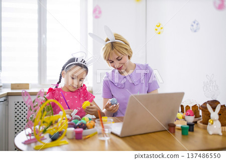 A mother and daughter wearing bunny ears paint Easter eggs together at a table while looking at a laptop screen, celebrating the holiday 137486550