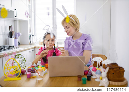 A mother and daughter wearing bunny ears participate in an online Easter egg decorating class, using a laptop for instruction and enjoying festive treats A mother and daughter wearing bunny ears participate in an online Easter egg decorating class, using a laptop for instruction and enjoying festive treats 137486554