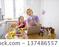 A mother and daughter wearing bunny ears are decorating Easter eggs together at a kitchen table with a laptop and festive treats 137486557