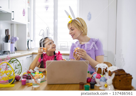 A mother and daughter wearing bunny ears are decorating Easter eggs together in a kitchen, with a laptop open on the table A mother and daughter wearing bunny ears are decorating Easter eggs together in a kitchen, with a laptop open on the table 137486561